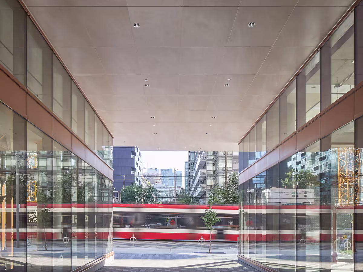 View from beneath a modern building's overhang framed by glass walls on both sides, showing a red and white streetcar passing by on the city street with tall buildings and trees in the background.