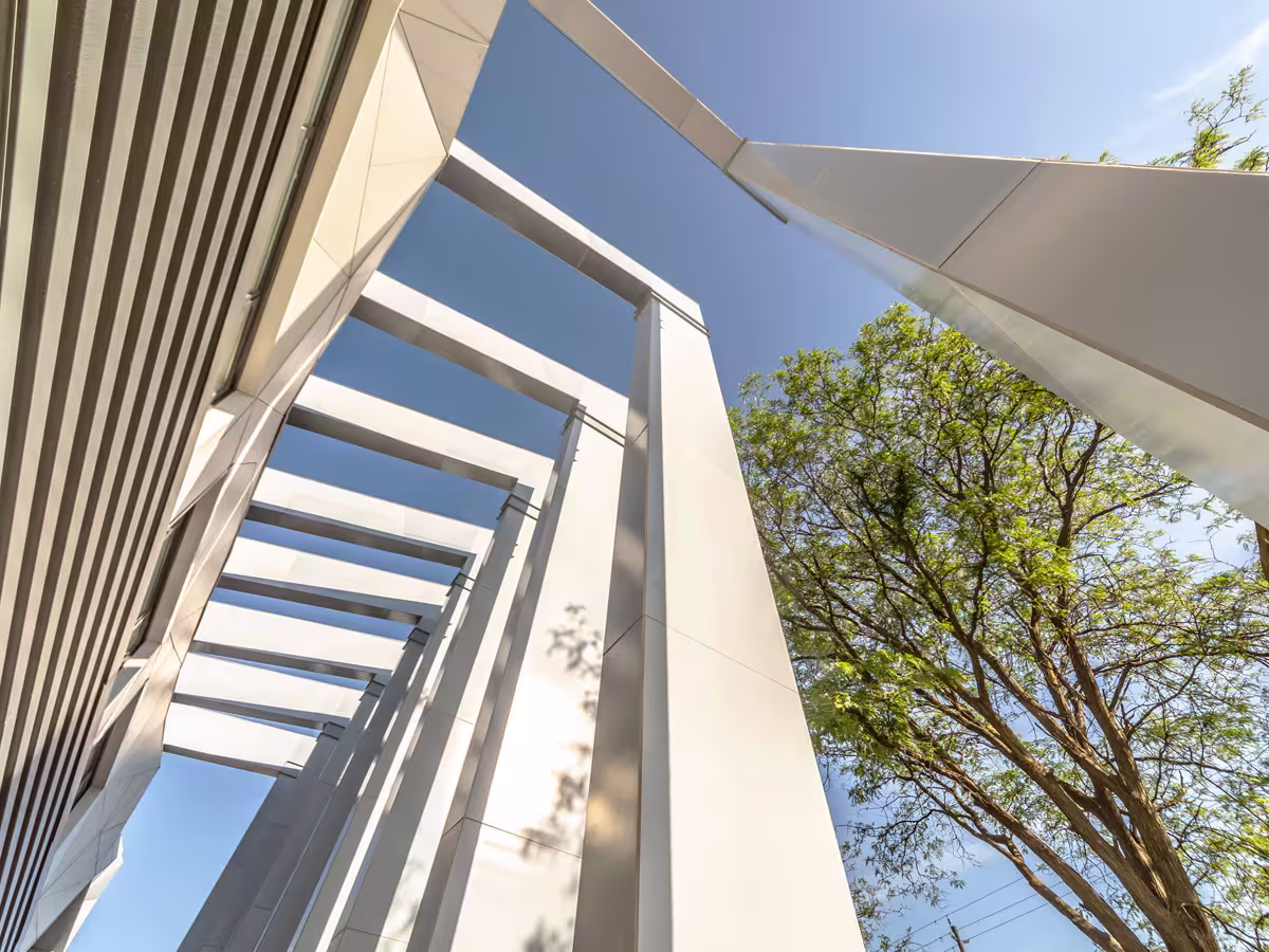 Upward perspective view of a modern architectural structure with tall white beams and a nearby green leafy tree against a clear blue sky.