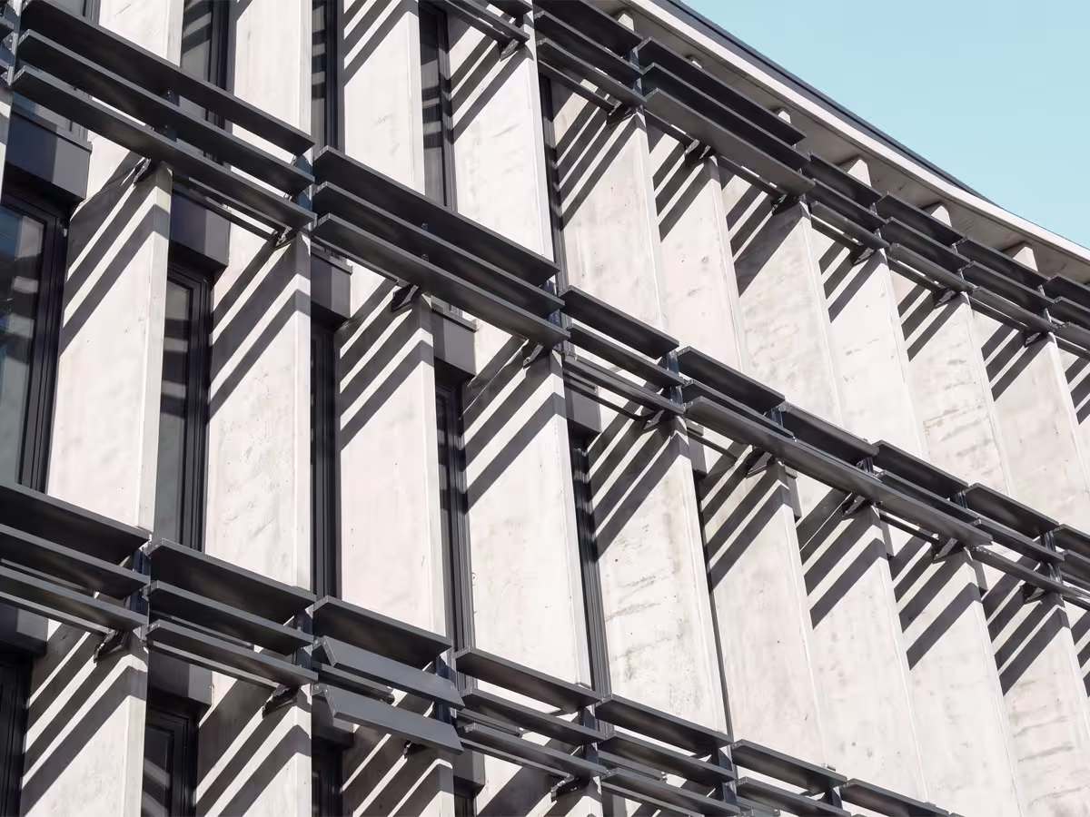 Close-up of a modern building facade featuring vertical concrete panels separated by narrow windows, with horizontal metal louvres casting diagonal shadows across the surface under a clear sky.