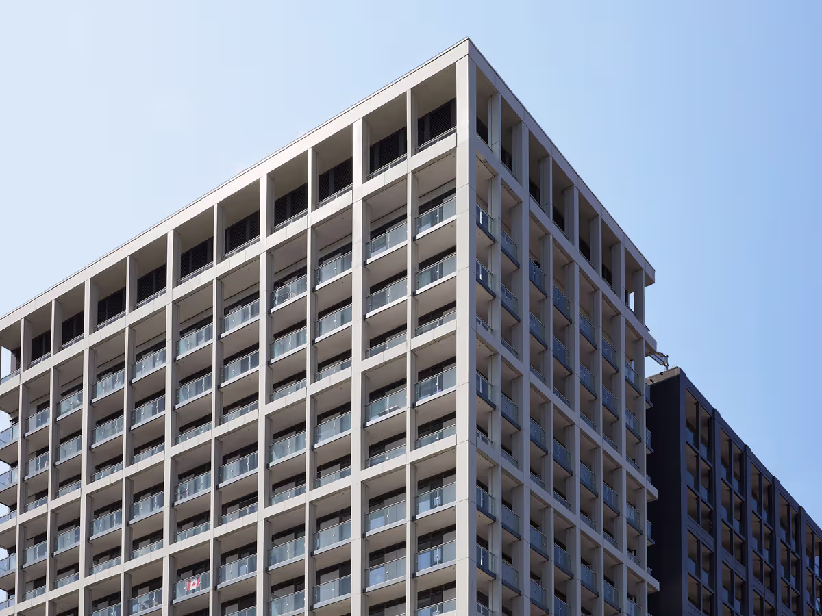 Modern high-rise residential building with multiple balconies featuring transparent railings under a clear blue sky.
