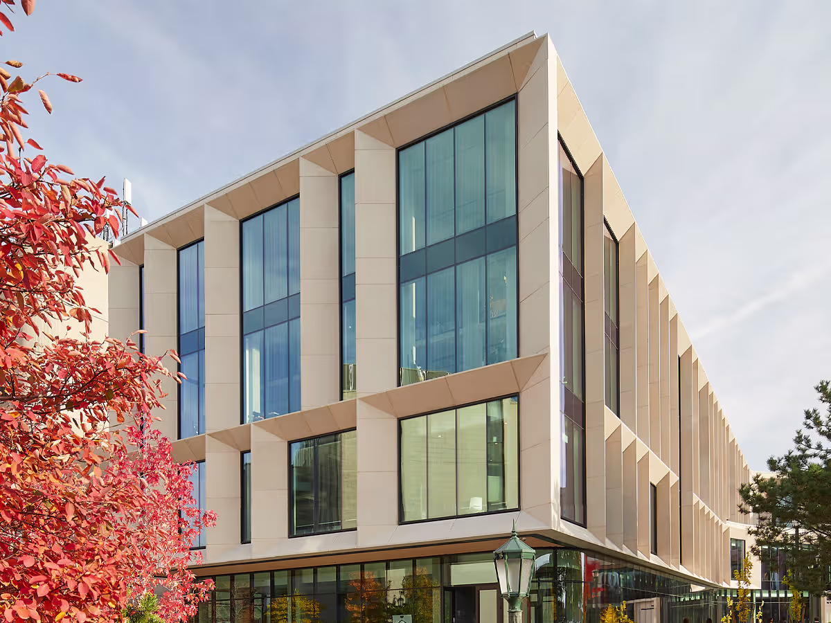 Modern beige office building with large vertical windows, surrounded by trees with red and green leaves under a clear sky.