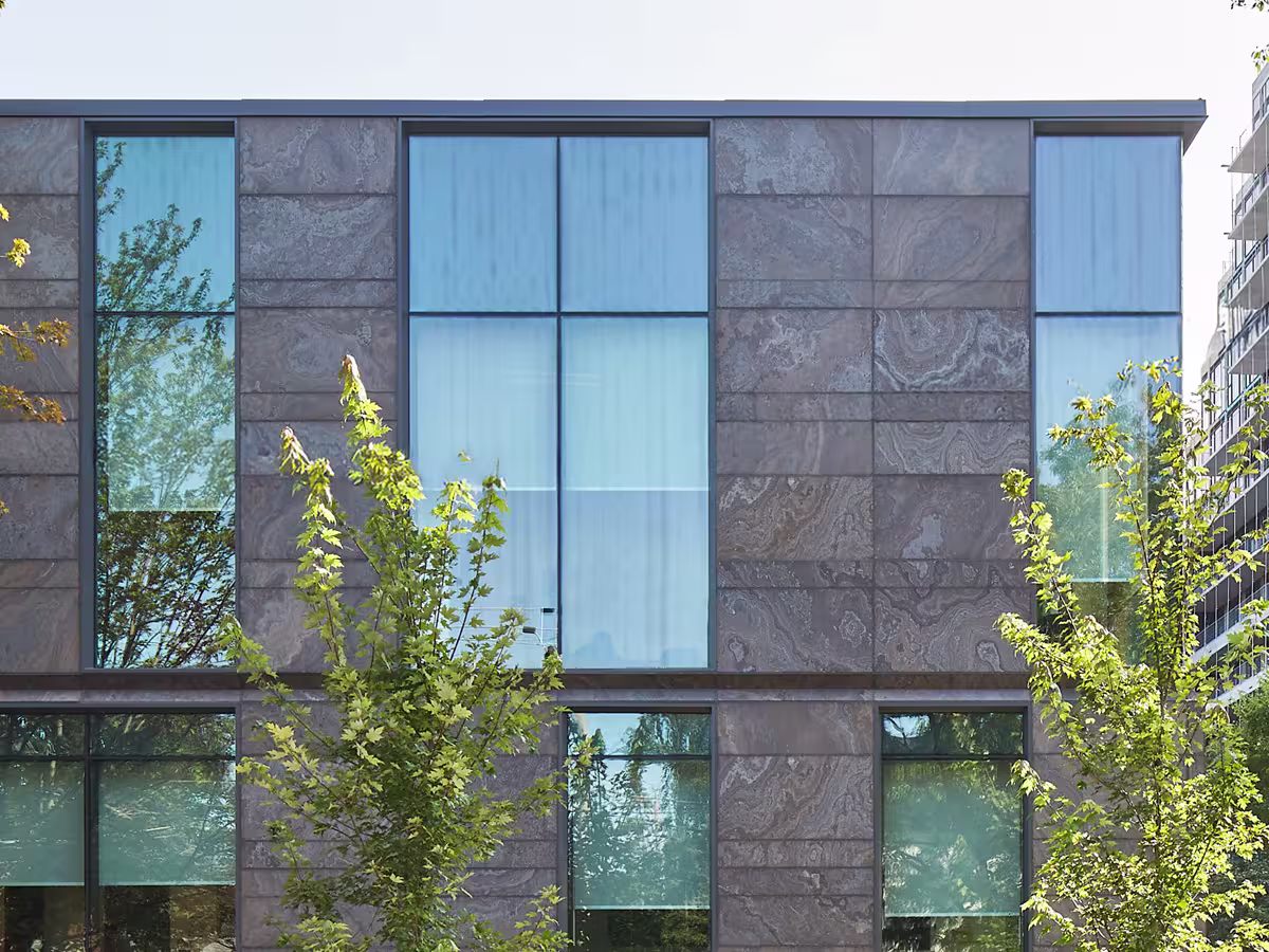 Modern building facade with large blue-tinted rectangular windows separated by stone panels and green leafy trees in front.