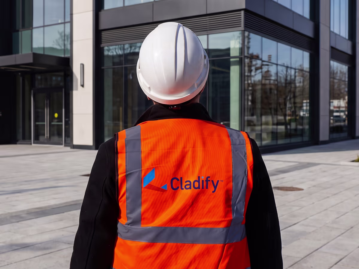 Person wearing a white construction helmet and an orange reflective safety vest with the blue Cladify logo on the back, standing outside a modern glass building.