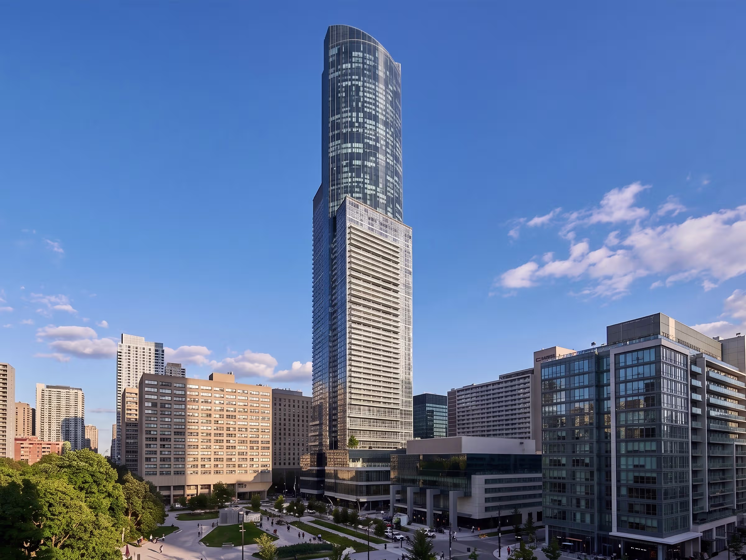 Tall modern skyscraper with glass facade surrounded by shorter buildings under a clear blue sky with some clouds, with a green space and people walking below.