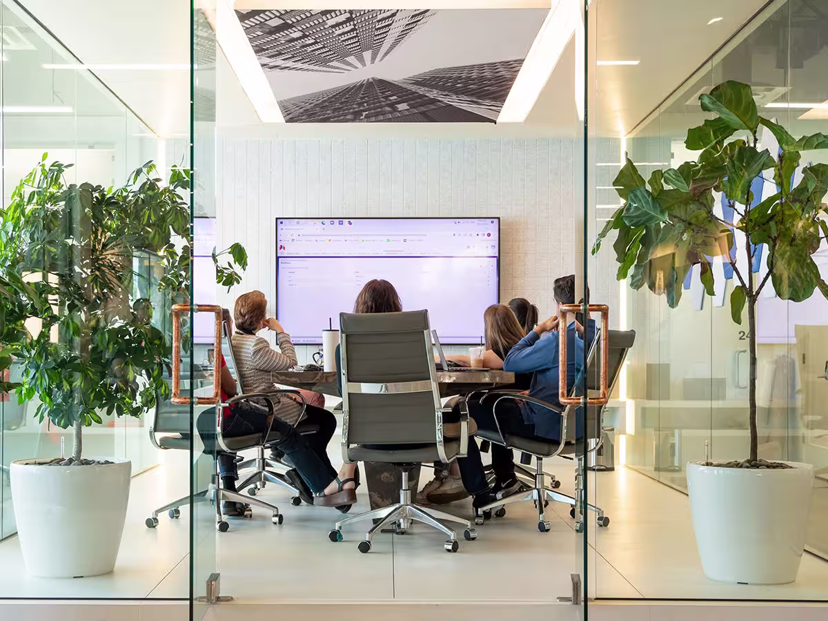 Four people sitting on office chairs around a conference table in a glass-walled meeting room, looking at a large screen displaying a computer interface, with large potted plants on either side of the entrance.