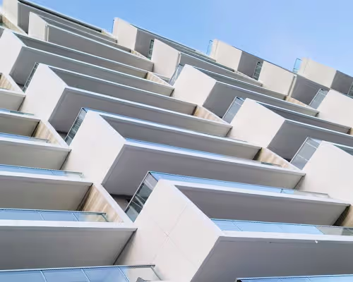Upward angled view of a modern building facade featuring a repetitive pattern of white rectangular balconies with glass railings against a clear blue sky.