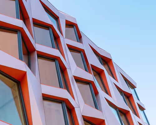 Modern building façade with a diagonal arrangement of large windows framed in orange and gray metal against a clear blue sky.