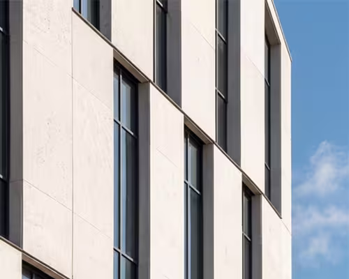 Close-up of a modern building facade with vertical tall windows and light-colored panels against a blue sky.