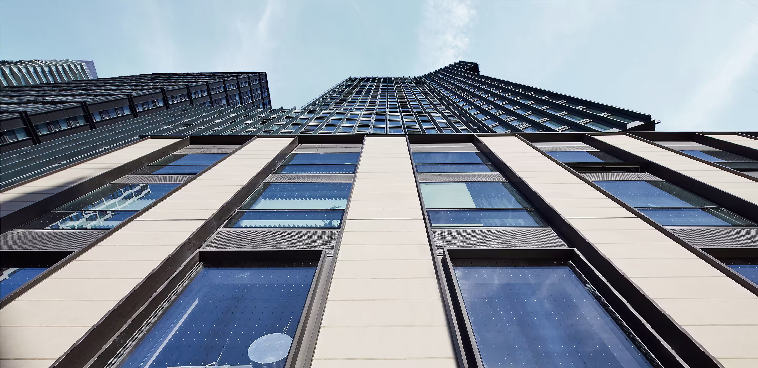 View looking up at a modern high-rise building with glass windows reflecting the sky and a nearby building visible on the left.