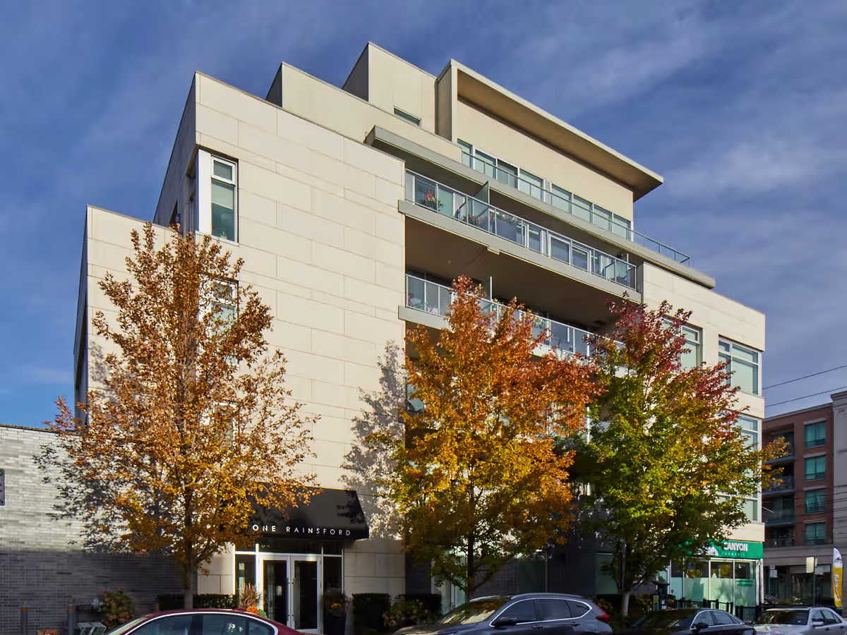 Multi-story beige building with balconies and large windows, partially obscured by three autumn trees with orange and green leaves, under a partly cloudy blue sky.