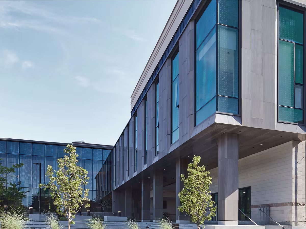 Modern building with large blue-tinted glass windows and stone columns under a clear sky, with young trees in front.