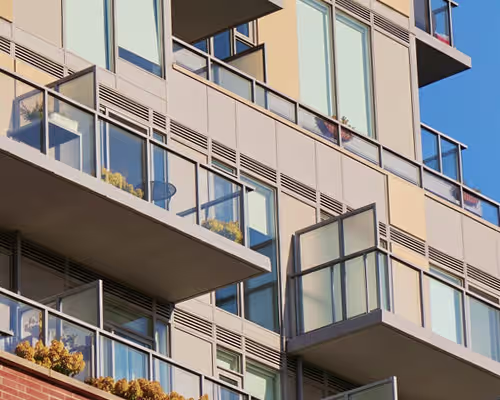 Modern building facade with multiple balconies featuring glass railings and some with open glass doors, under a clear blue sky.