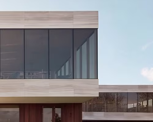Modern building exterior featuring large dark tinted floor-to-ceiling windows framed by light wood paneling under a clear blue sky.
