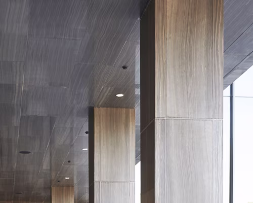 Interior architectural detail showing modern vertical columns and ceiling on a building, featuring a smooth grey ceiling and beige stone-textured columns with natural light coming through glass windows.