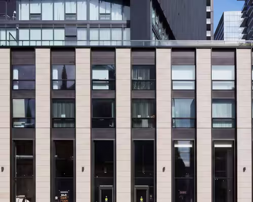 Modern building facade with large vertical windows framed by dark panels and beige stone columns, reflecting surrounding structures.