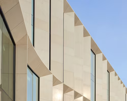 Close-up of modern building facade featuring tan-colored panels with geometric shapes and vertical windows against a clear blue sky.