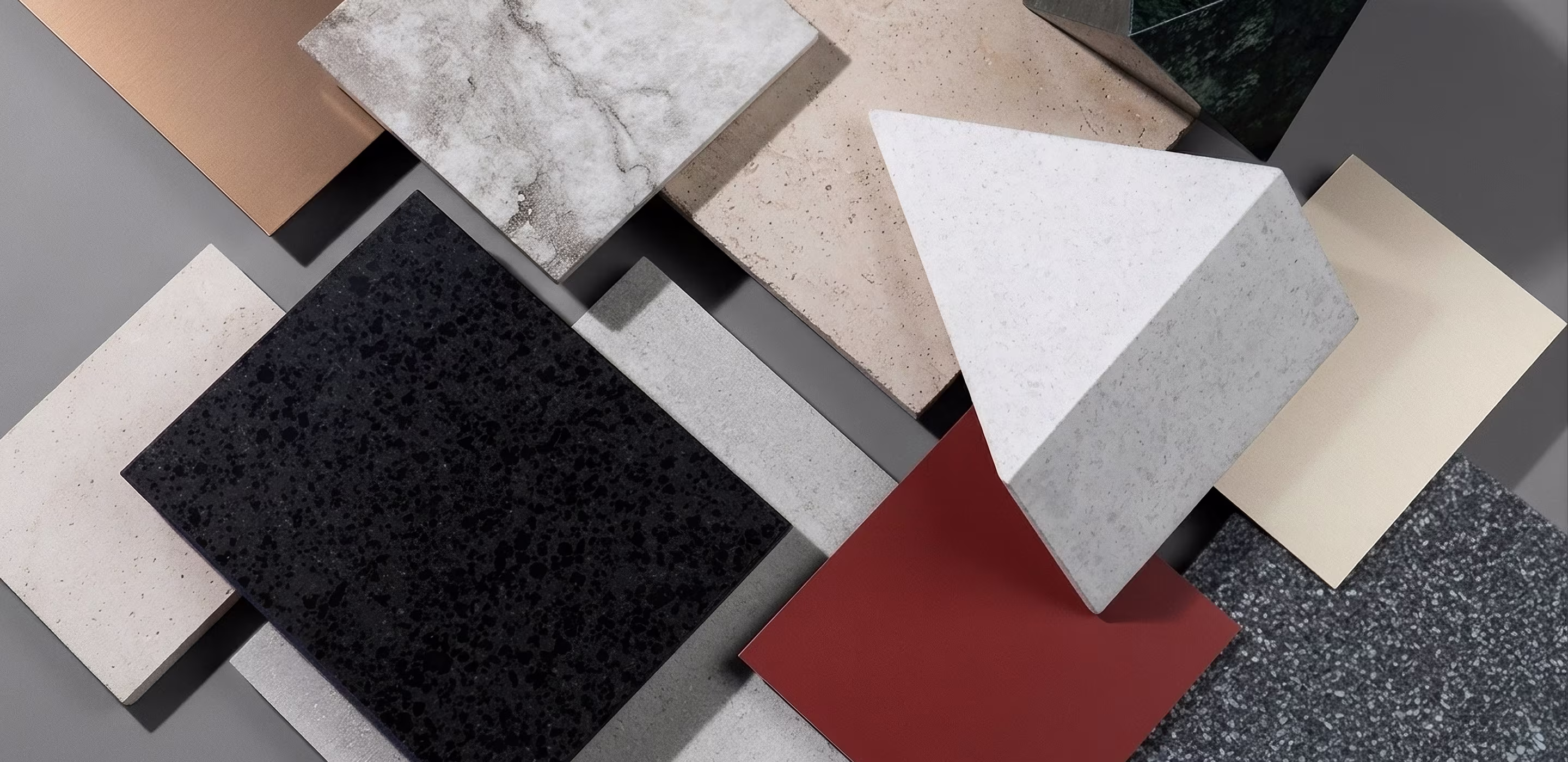 Various ceramic and stone tiles in different colors and textures arranged on a gray surface, including white, black, brown, beige, terracotta, gray, and speckled patterns.