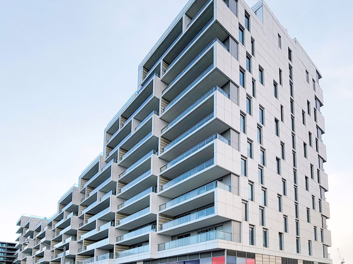 Modern multi-story building with staggered balconies covered with glass railings, light-colored exterior panels, and multiple windows under a clear sky.