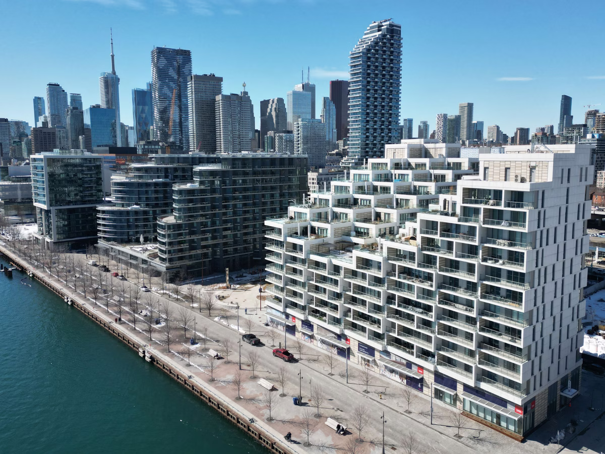 Modern stepped condominium building along a waterfront promenade, with a city skyline of high-rise buildings in the background under a clear blue sky.
