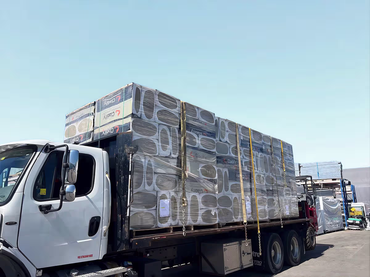 White flatbed truck loaded with multiple large stacks of boxed materials wrapped in plastic secured by yellow straps under clear blue sky.