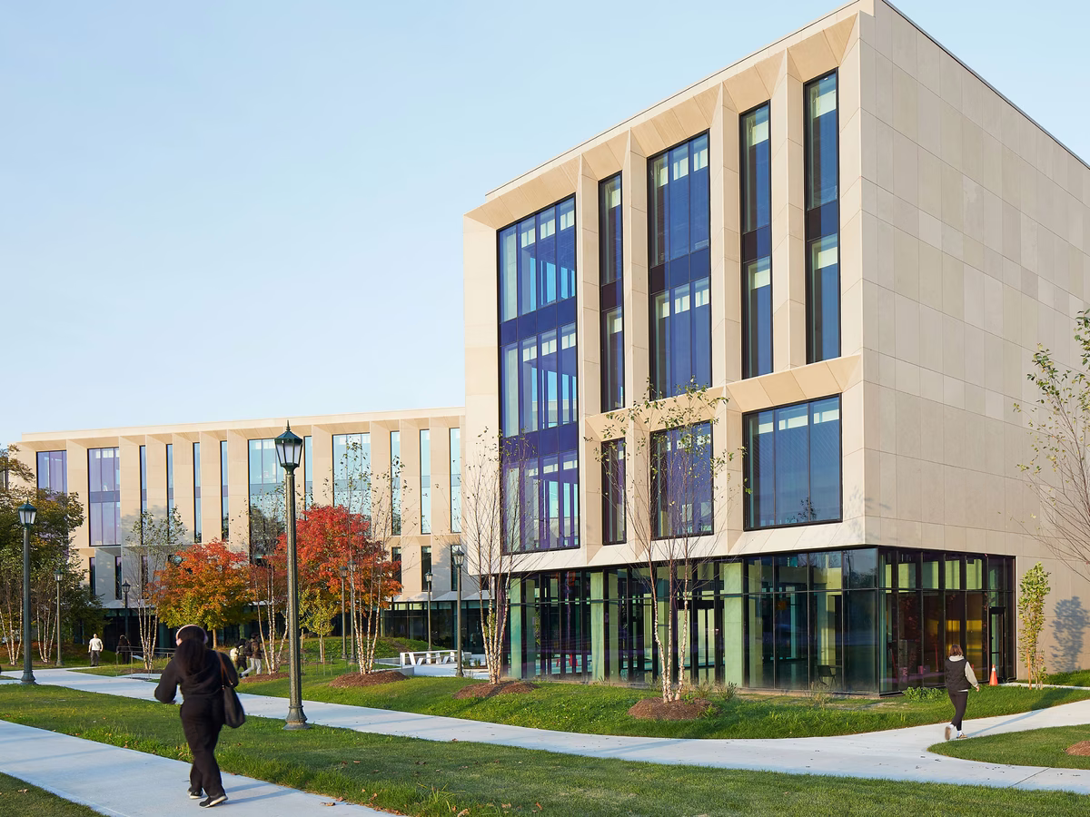 Modern university campus building with large vertical windows, surrounded by walkways, green lawns, trees with autumn foliage, and two people walking.