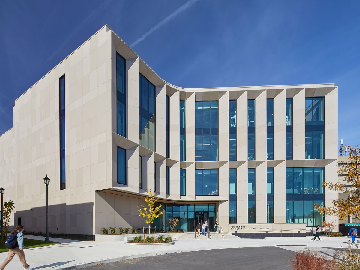 Modern university building with large vertical windows and beige facade under a clear blue sky with a contrail, with a few people walking nearby.