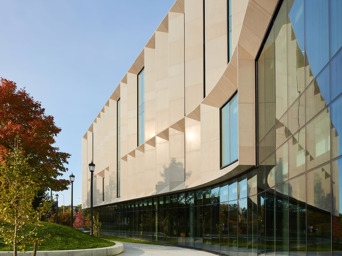Modern building with light beige facade and large glass windows reflecting surrounding trees, under a clear blue sky during daytime.