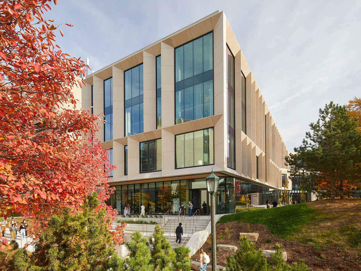 Modern university building with large vertical windows, surrounded by autumn trees with red and orange leaves, and people walking near stairways and pathways.