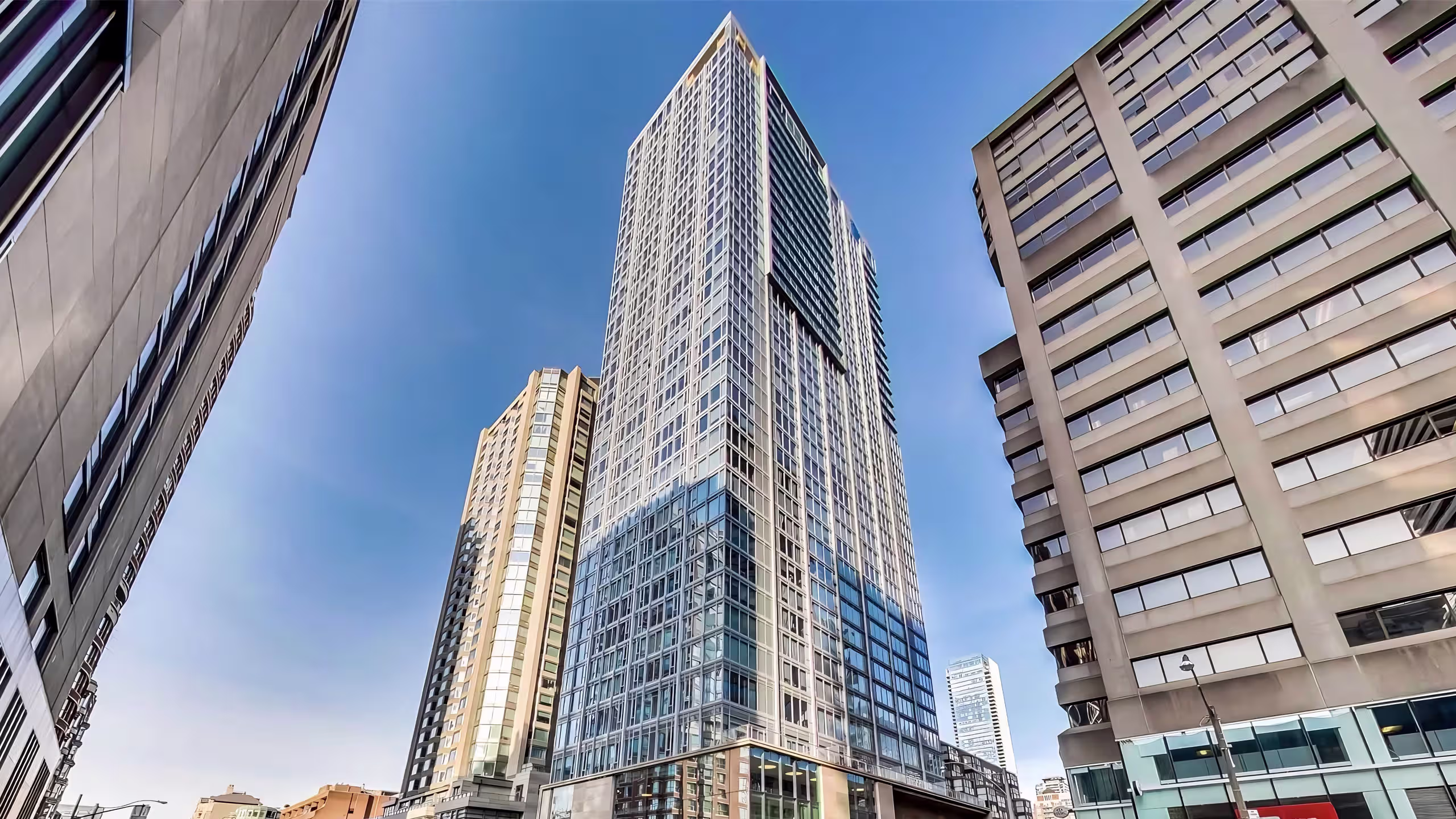 Low-angle view of a modern tall glass and steel skyscraper surrounded by other high-rise buildings under a clear blue sky.
