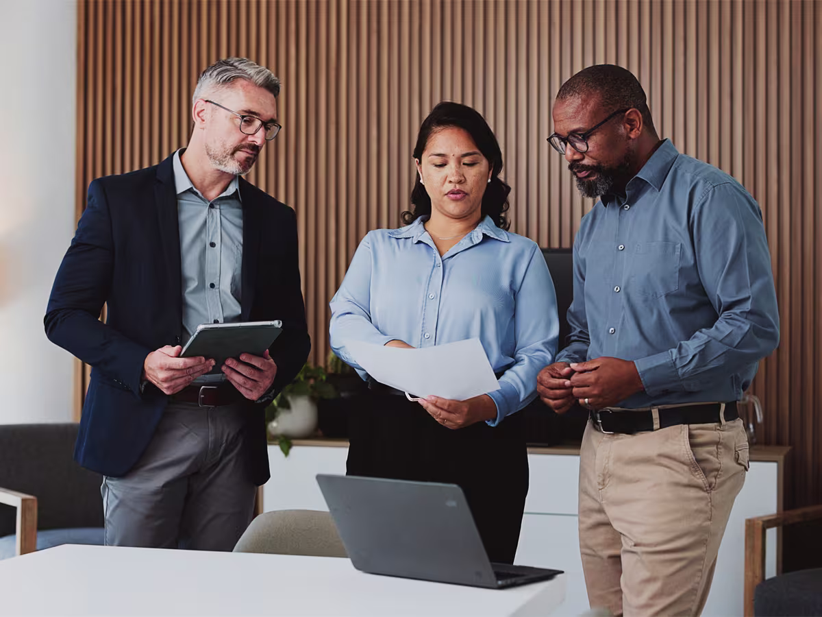 Three professionals, two men and one woman, standing indoors around a table with a laptop; the woman in the center holds a document while the men look at it, engaged in discussion.