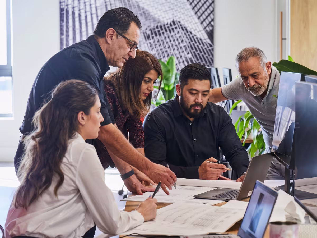 Five diverse professionals collaborate over architectural blueprints and laptops in a modern office, discussing design plans.