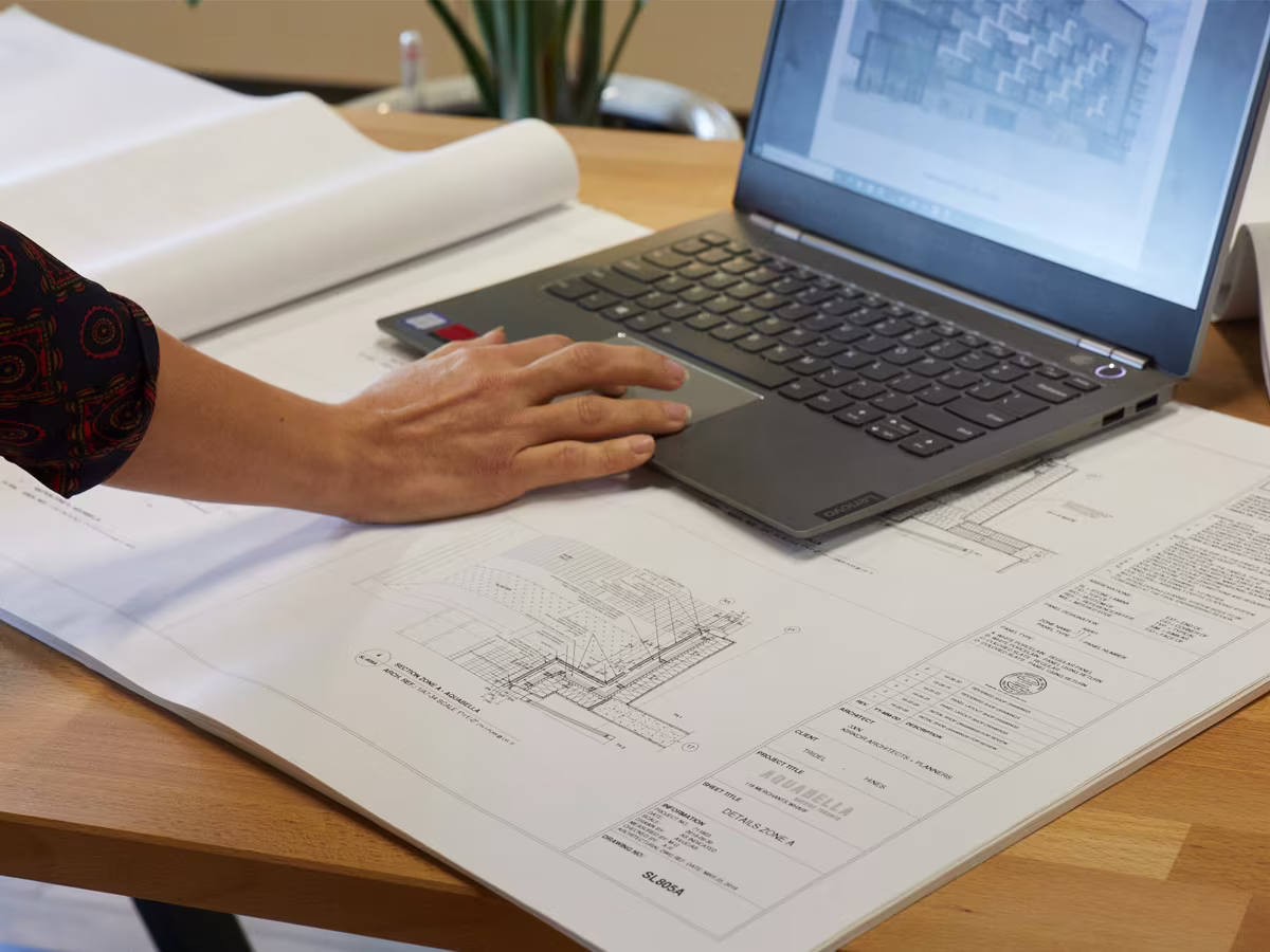 A person's hand using a laptop placed on top of architectural blueprints on a wooden table, with technical drawings displayed on the laptop screen.