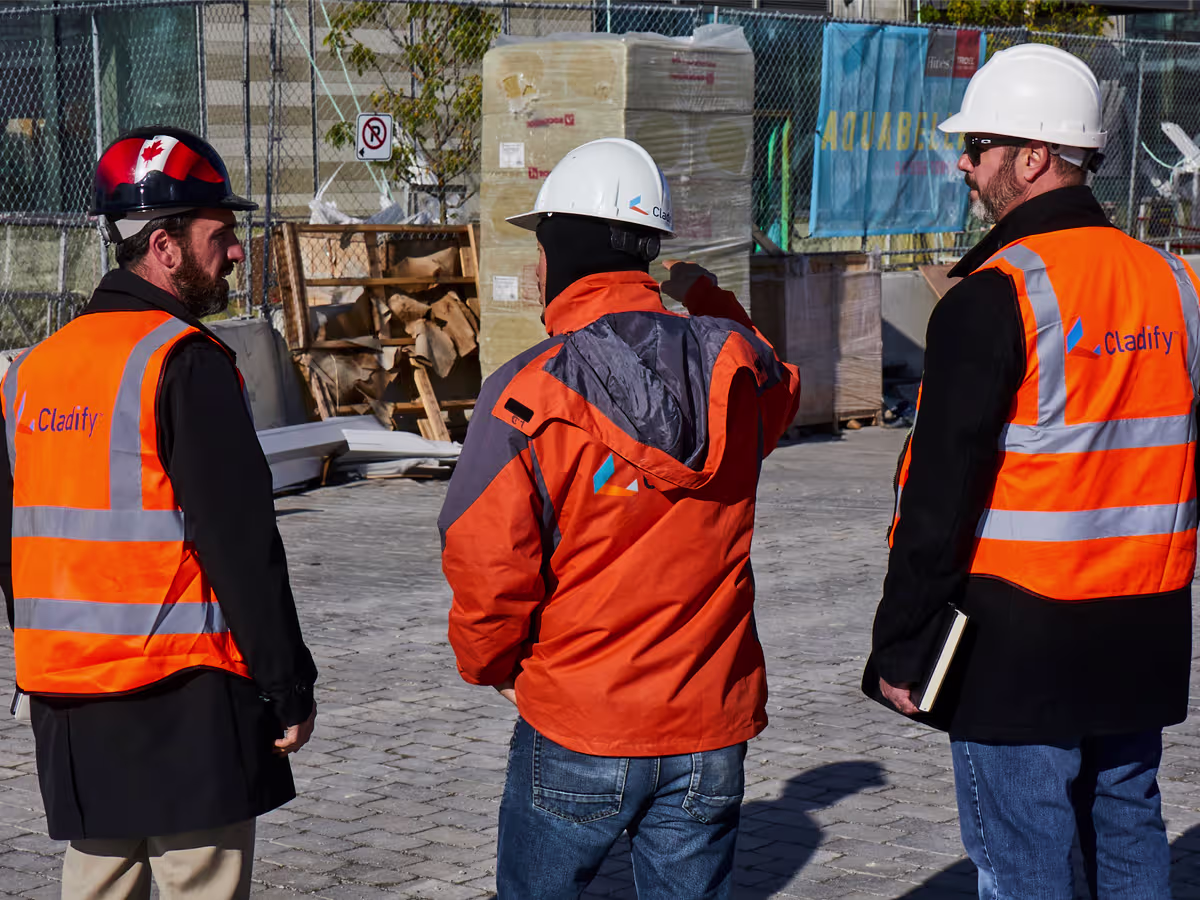 Three construction workers wearing safety helmets and bright orange vests with the logo 'Cladify' stand on a paved site, appearing to discuss or inspect the area with construction materials and fencing in the background.