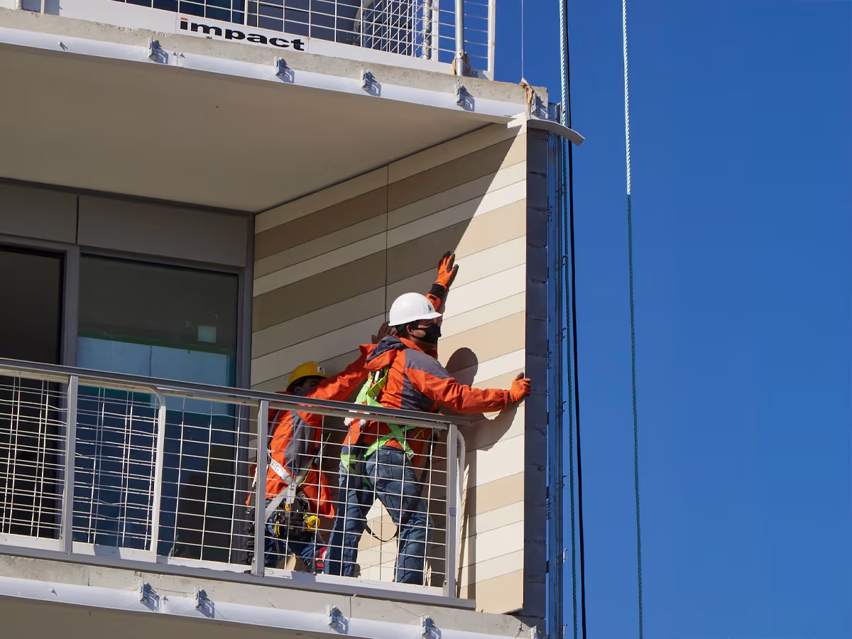 Two construction workers wearing hard hats and safety gear installing or inspecting vertical paneling on the exterior corner of a modern building balcony under clear blue sky.