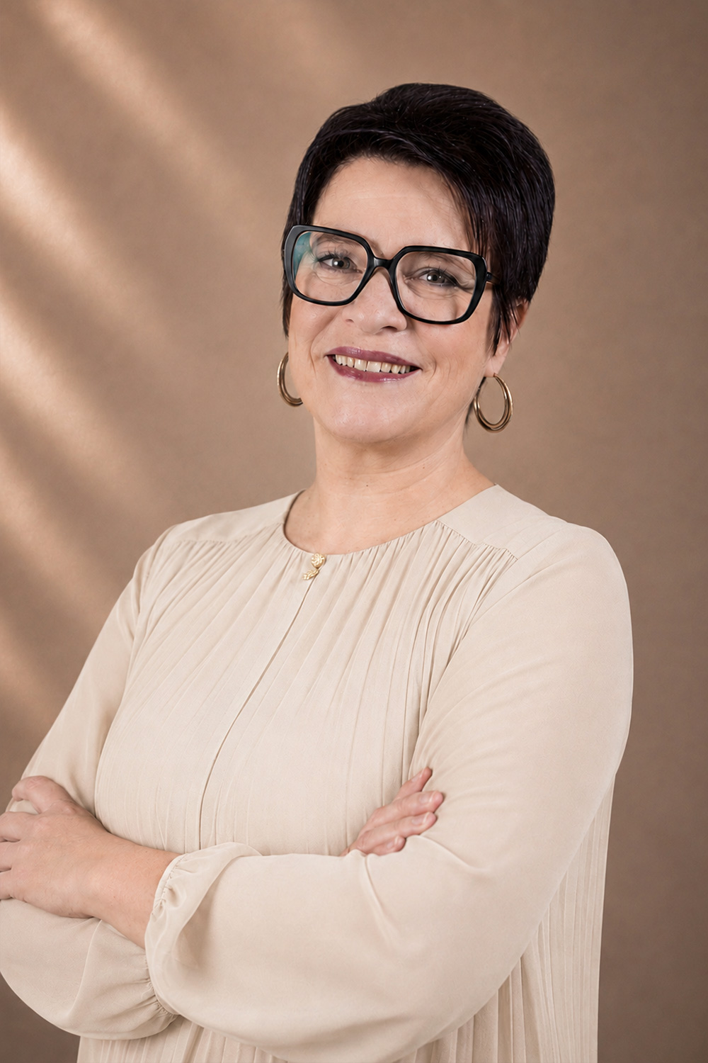 Frau mit kurzen schwarzen Haaren, großer Brille und beiger Bluse lächelnd mit verschränkten Armen.