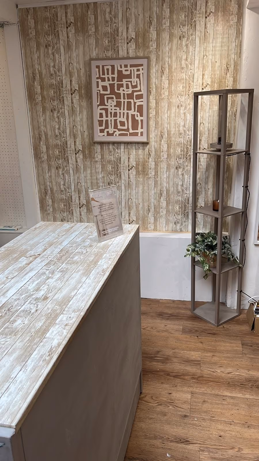 Interior corner of a room with rustic wooden plank walls and floor, a light-colored wooden counter in the foreground, abstract geometric artwork on the wall, and a tall narrow shelving unit holding plants and small items.