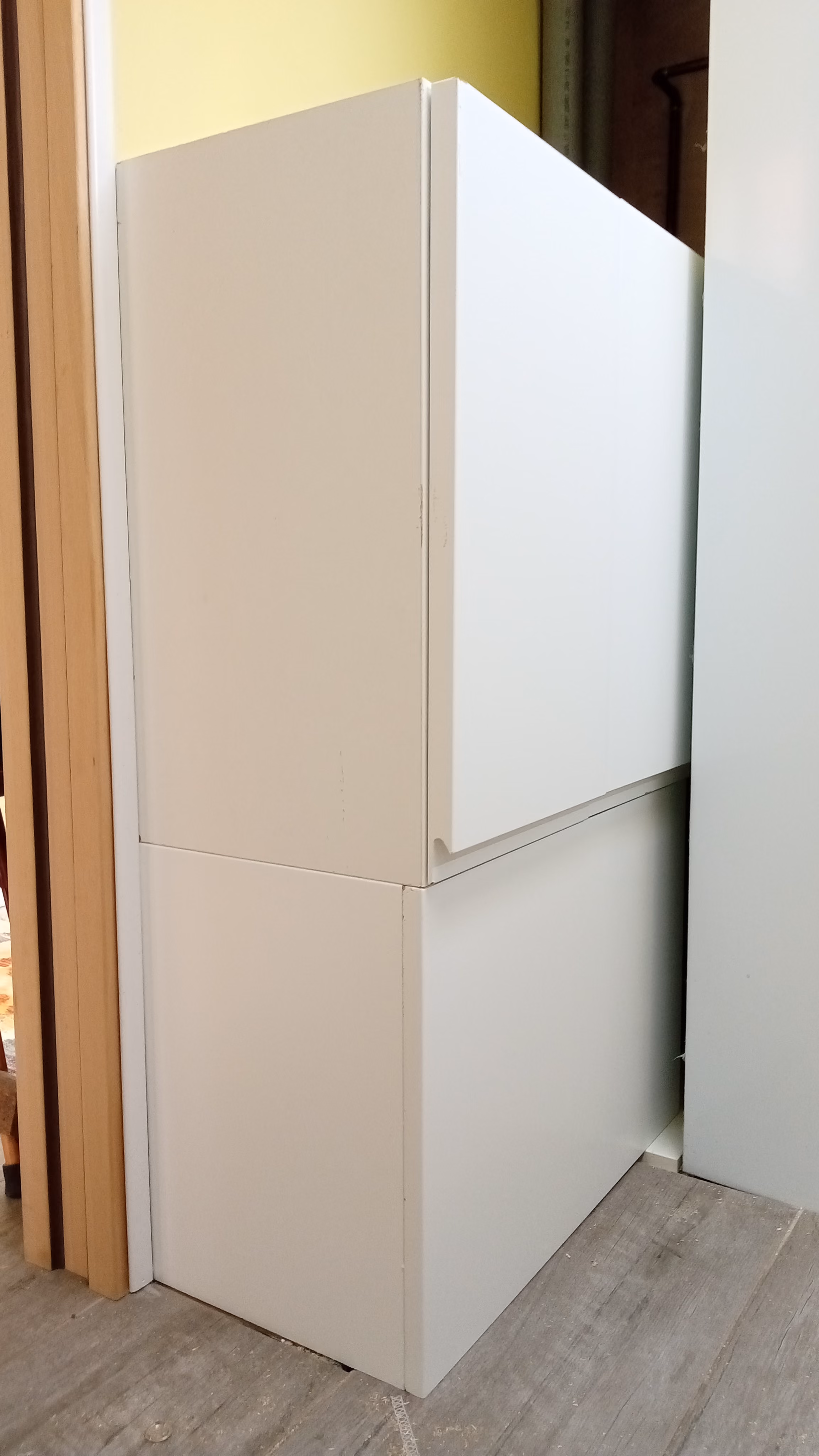 Two stacked white cabinets positioned in a corner, next to a wooden door frame on the left and a grayish wall on the right, with a light-colored floor beneath.