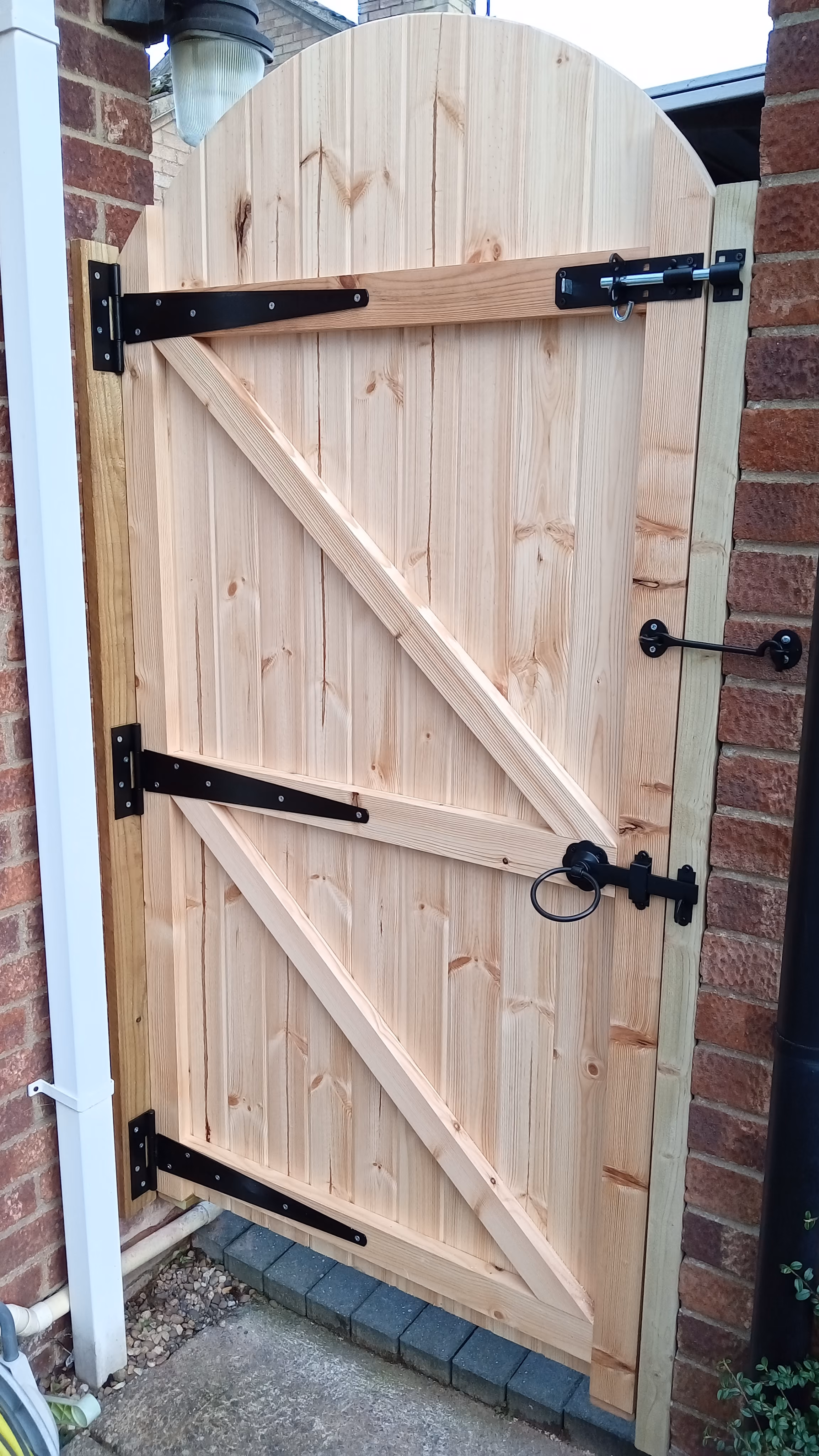 Light natural wooden garden gate with black metal hinges and latch, installed between brick and wooden posts.