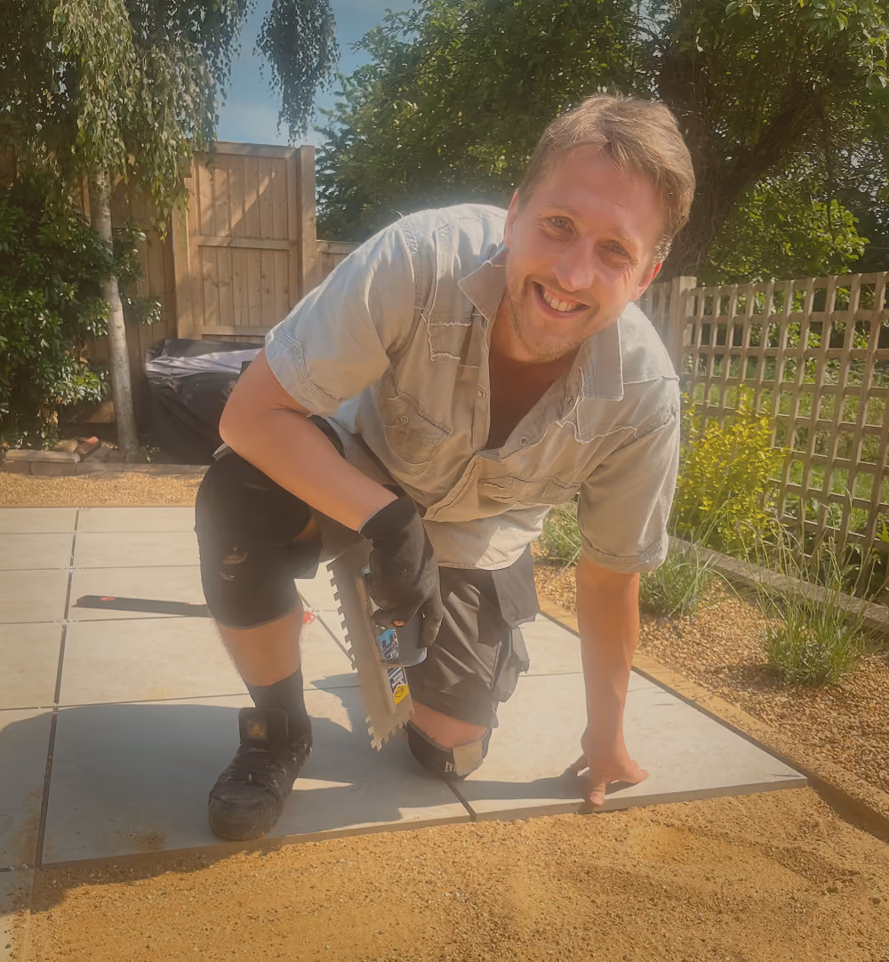 Ray smiling and kneeling on outdoor tiles while holding a notched trowel, surrounded by garden plants and a wooden fence.