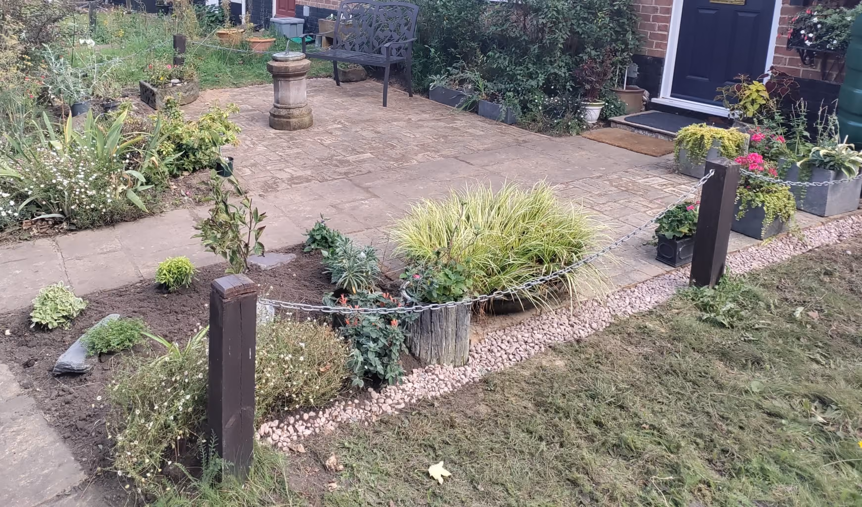 Small garden area bordered by a wooden post and chain fence with various green plants and flowers, adjacent to a paved patio with a bench and a birdbath near a brick house with a black door.