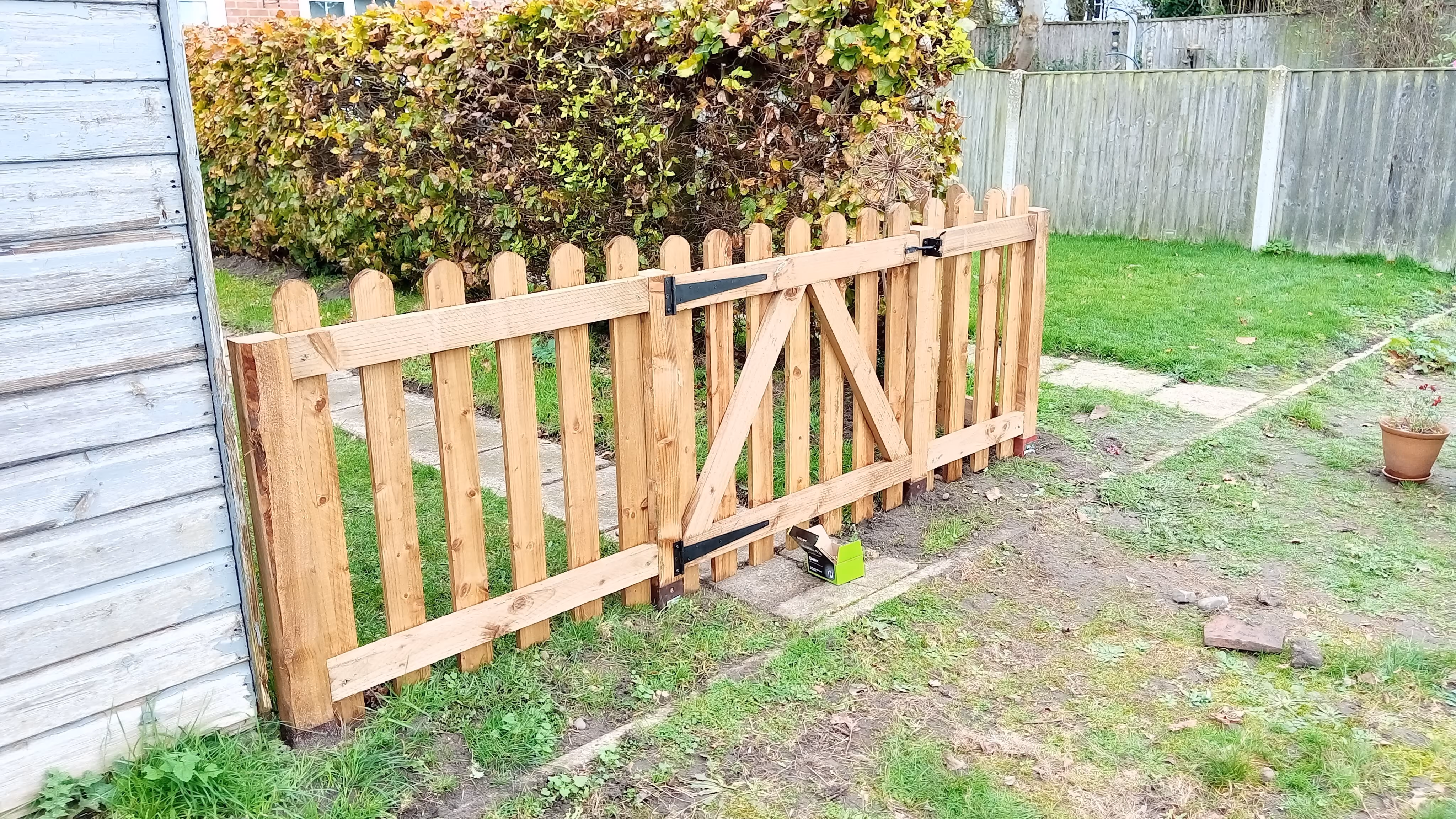 A small wooden garden gate attached to a wooden fence with a latch, set in a grassy backyard with a hedge and a weathered wooden building on the left.