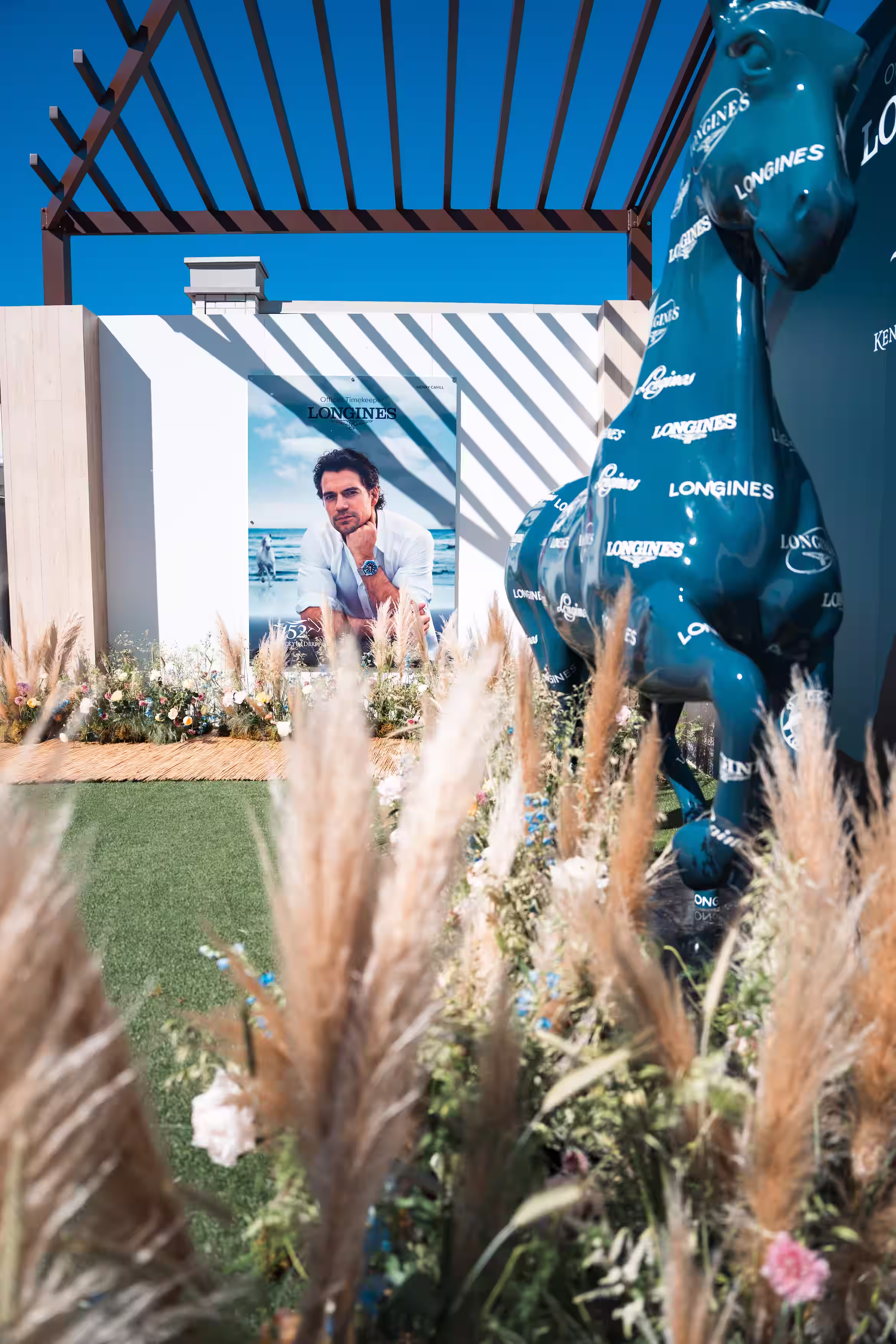 Outdoor scene with Longines branded blue horse statues, plants, and a poster of a man wearing a watch.