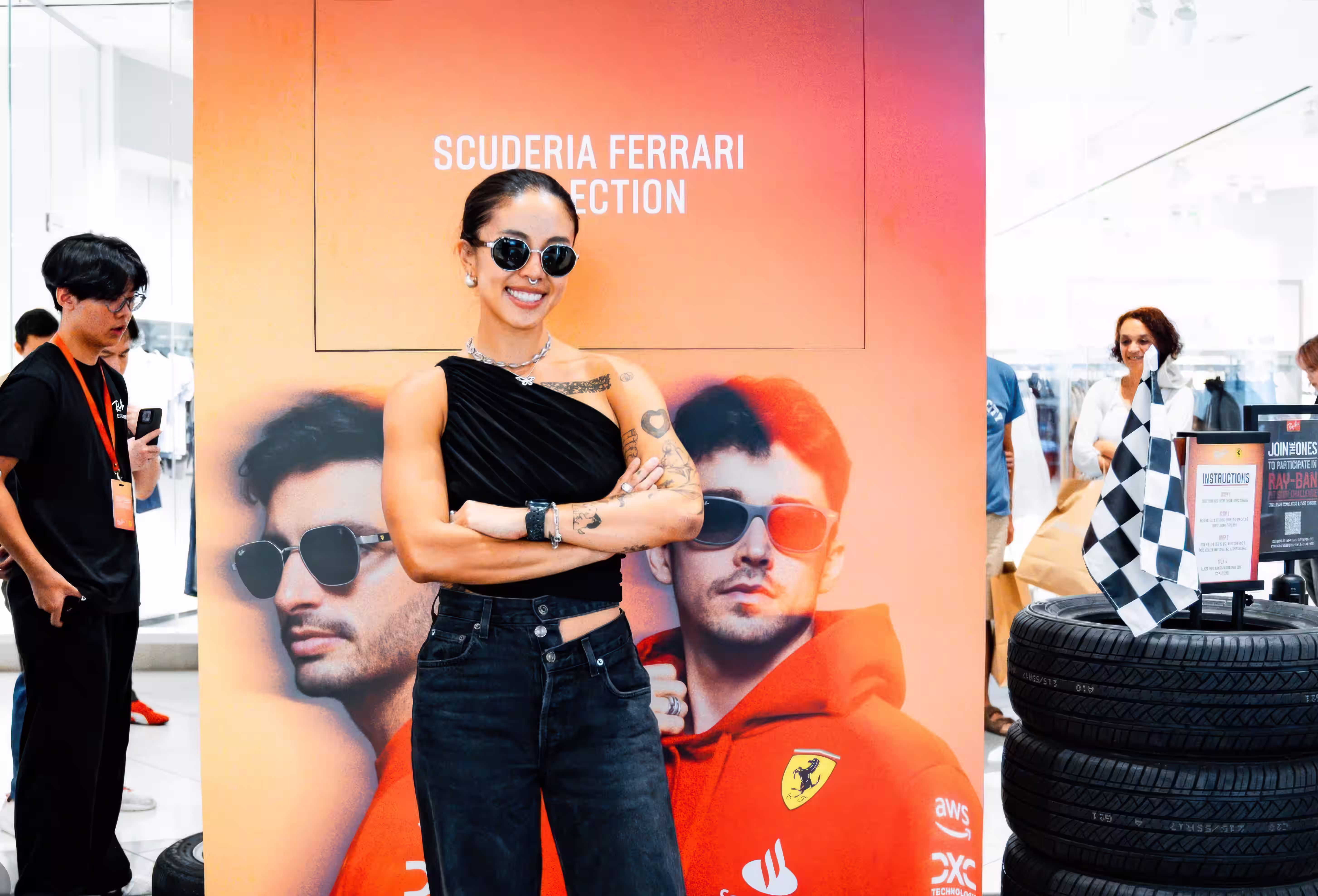 Smiling woman with tattoos and sunglasses poses in front of a Scuderia Ferrari Collection poster.