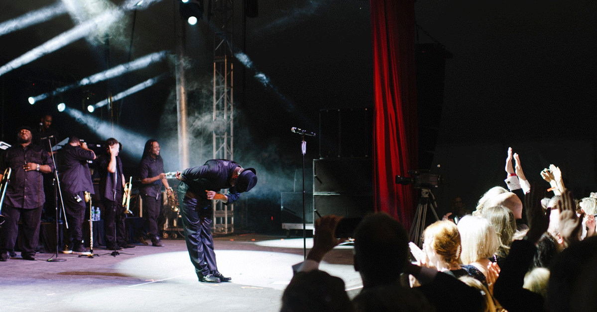 Performer dressed in black bowing on stage while band members stand and audience claps and cheers.