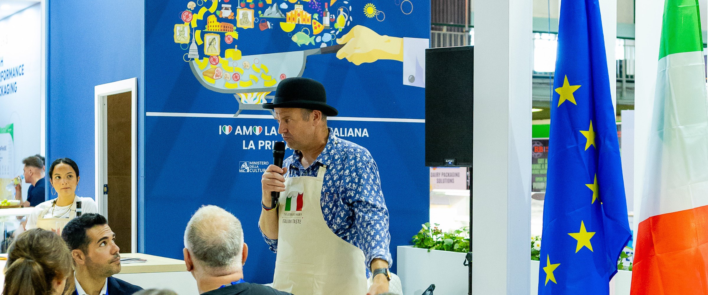 A presenter at an international pavilion with EU and Italian flags