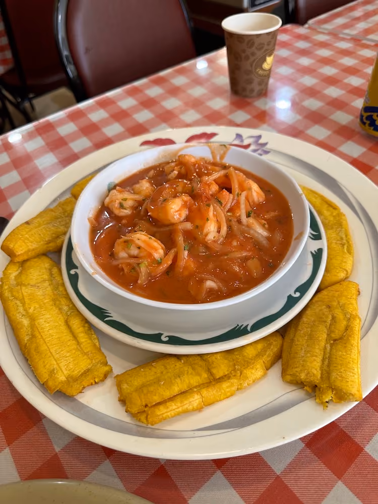 Bowl of shrimp ceviche in red sauce with fried plantain slices on a white plate on a red checkered tablecloth.