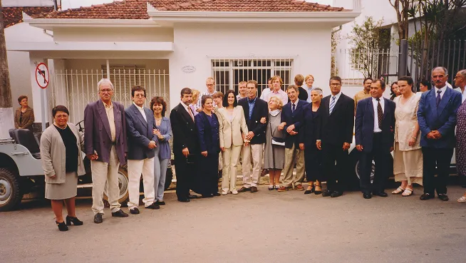 A large group of people dressed formally standing outdoors in front of a white house with a tiled roof.