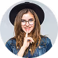 Young woman with glasses smiling and holding her finger to her lips in a shushing gesture.