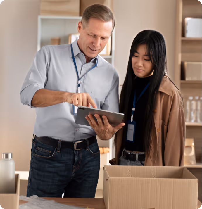 Two people with ID badges looking at a tablet, standing near an open cardboard box in a room with shelves.