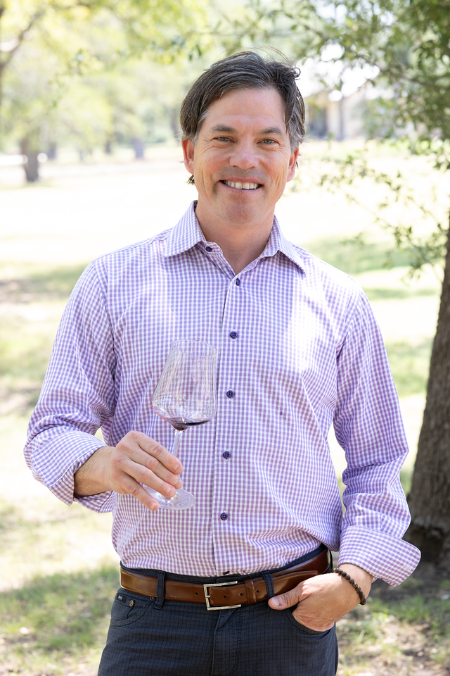 A man smiling outdoors holding a glass of red wine, dressed in a light purple checkered shirt and dark pants, standing in a sunlit park with trees in the background.